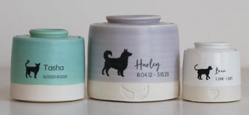 Three ceramic pet memorial urns in different sizes, featuring a pastel gradient design and black silhouette pet illustrations with personalized names and dates.