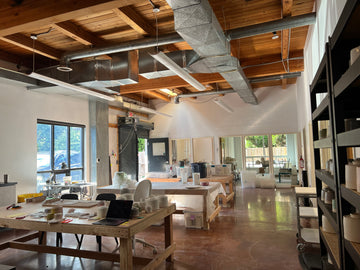 A wide interior view of our pottery studio or workshop with wooden tables, ceramic vessels, shelves, large windows, exposed wood ceiling beams, and metal ductwork. No single retail product is isolated; the scene itself is the main visual content.